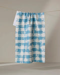 The Geometry Patch Denim recycled kitchen towel, featuring a blue and white checkered textured pattern, hangs on a clothesline against a neutral background.