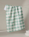 The Geometry Patch Mint tea towel, featuring a green and white checkered pattern, hangs on a line with sunlight casting soft shadows. Stacked white ceramic dishes appear in the lower right corner.