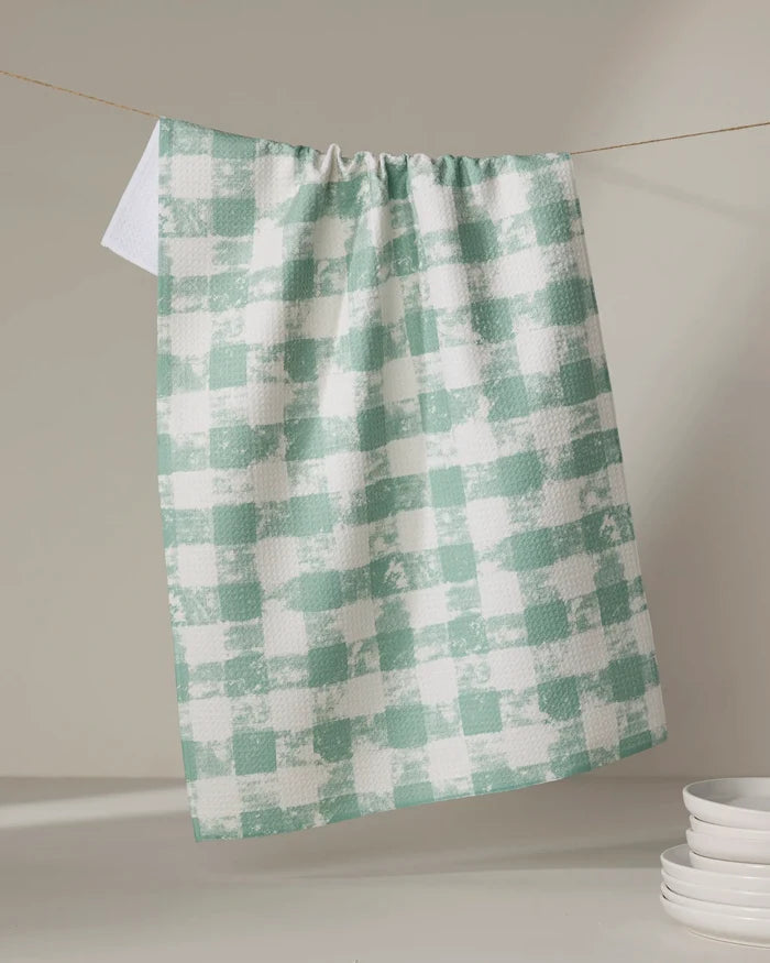 The Geometry Patch Mint tea towel, featuring a green and white checkered pattern, hangs on a line with sunlight casting soft shadows. Stacked white ceramic dishes appear in the lower right corner.