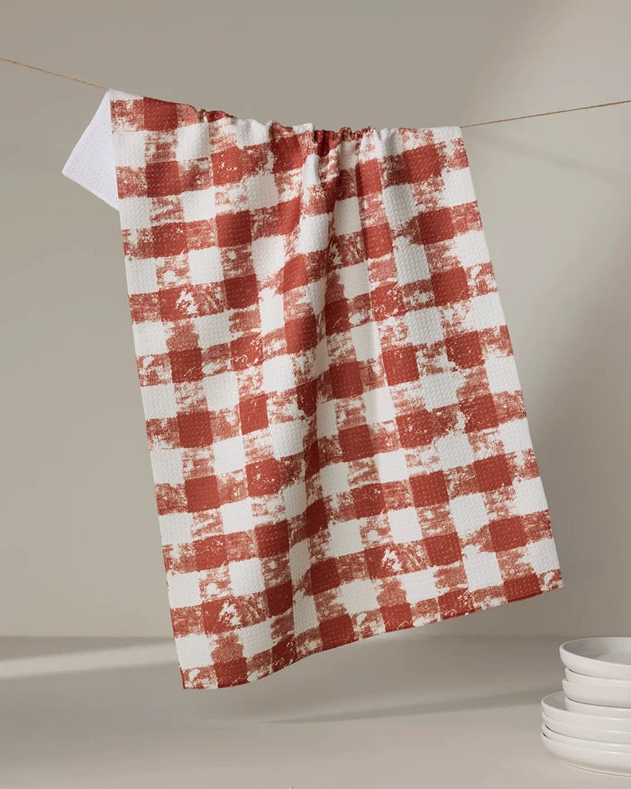 The Geometry Patch Spice tea towel, featuring a red and white checkered design, hangs on a string against a neutral background, with a stack of white plates partially visible in the lower right corner.