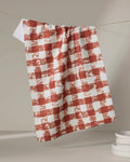 The Geometry Patch Spice tea towel, featuring a red and white checkered design, hangs on a string against a neutral background, with a stack of white plates partially visible in the lower right corner.
