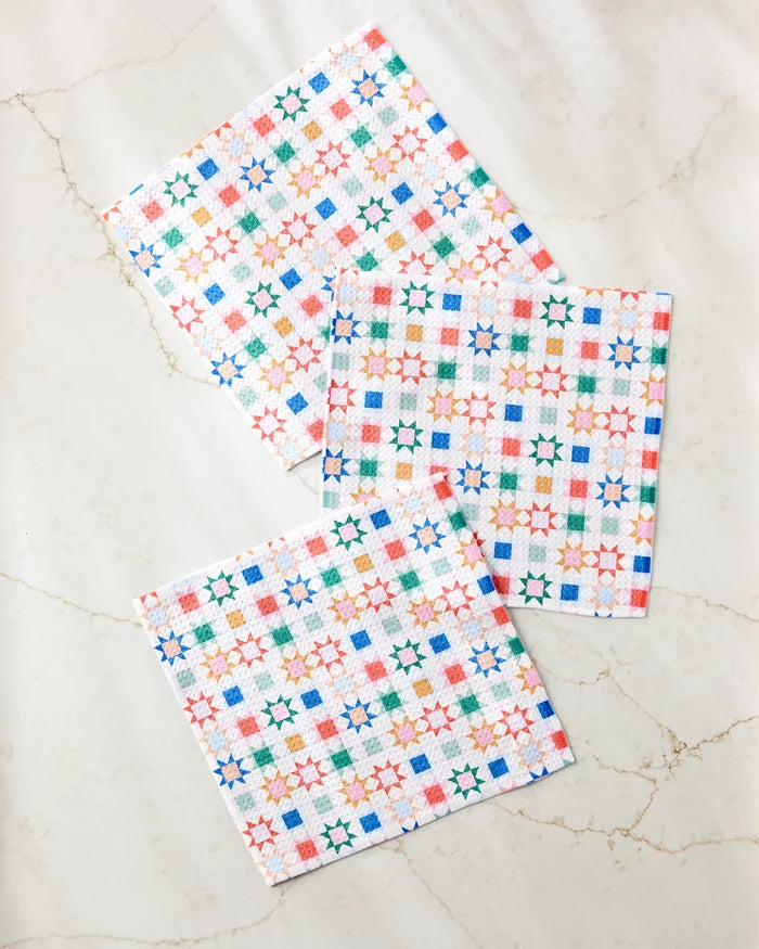 The Patchwork Picnic dishcloth set by Brittany Jepsen features three square cloths with vibrant geometric star and square patterns in orange, green, blue, and red—made from recycled materials and displayed on a marble-like surface.