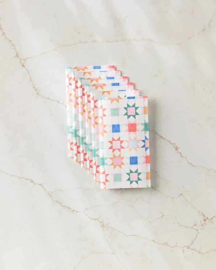 Six Patchwork Picnic kitchen towels by Brittany Jepsen, featuring a colorful geometric star pattern, are neatly folded and stacked on a light marble surface.