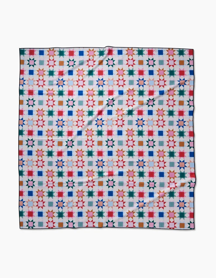 Patchwork Picnic by Brittany Jepsen is a square beach blanket crafted from recycled materials, showcasing a vibrant geometric star pattern in red, blue, green, and pink on white. The fabric is slightly wrinkled and rests flat on a plain surface.