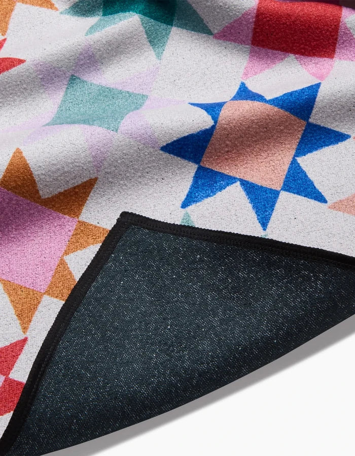 A close-up of the Patchwork Picnic beach blanket by Brittany Jepsen, showcasing its recycled material and colorful geometric star pattern. One corner is folded to reveal the dark, textured underside.