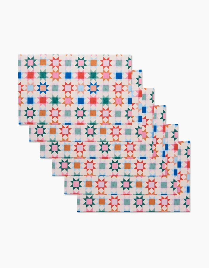 The Patchwork Picnic placemats by Brittany Jepsen feature a geometric star design in red, blue, pink, green, and yellow—crafted from eco-friendly post-consumer recycled materials—shown stacked on a white background.