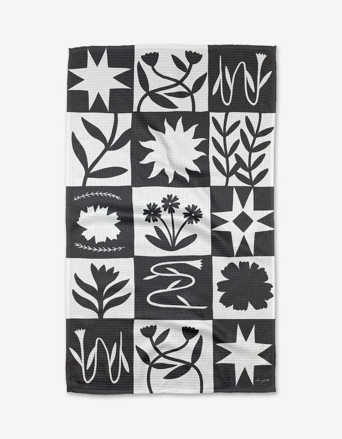 The Patchwork Tea Towel by Elana Gabrielle features a black and white grid of stylized flowers, stars, and bold lines. Made from recycled materials, its graphic symmetry brings modern flair to any kitchen.