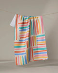 The Pathways tea towel by Sareka Unique features vibrant pink, orange, blue, and green brushstrokes in a grid pattern and hangs on a string against a neutral background.