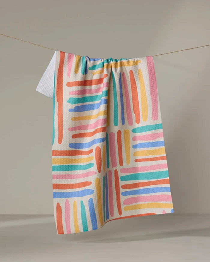 The Pathways tea towel by Sareka Unique features vibrant pink, orange, blue, and green brushstrokes in a grid pattern and hangs on a string against a neutral background.