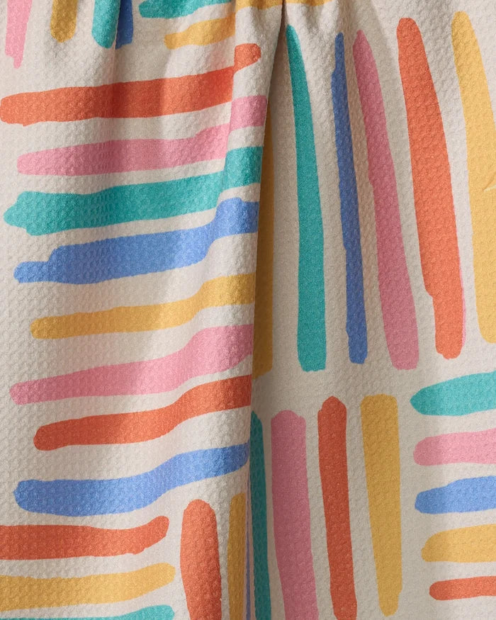 Close-up of Sareka Unique's Pathways tea towel, showcasing textured fabric with uneven, colorful stripes in orange, yellow, pink, blue, and teal on a light beige background. Visible seam and gathered section at the top add detail.