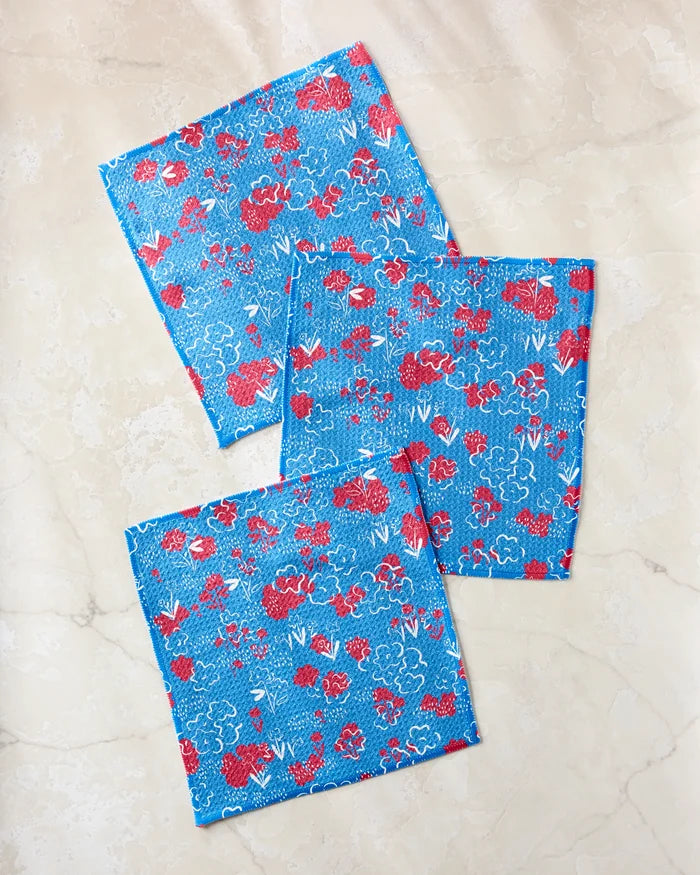 Three blue cloth napkins with red and white floral designs from Second Nature Studio’s Patriotic Garden set are arranged on a light marble-like surface.