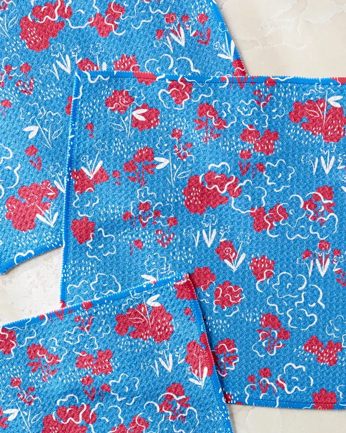 Four blue “Patriotic Garden” dishcloths by Second Nature Studio, featuring red and white floral patterns with stylized flowers and leaves, are arranged on a beige, marble-like surface.