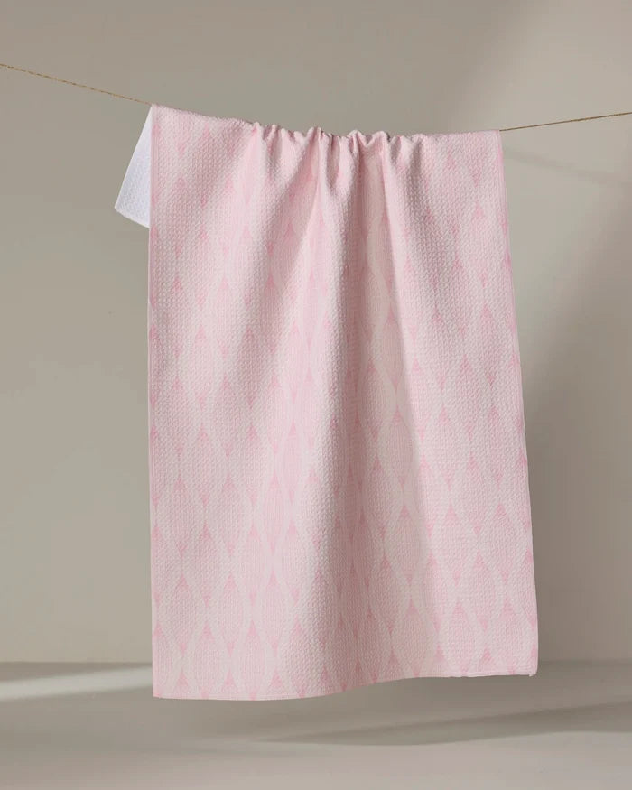 The Shelley Smith "Patterned in Pink" kitchen towel, made from recycled materials with a subtle diamond design, hangs neatly on a clothesline against a neutral background.