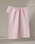 The Shelley Smith "Patterned in Pink" kitchen towel, made from recycled materials with a subtle diamond design, hangs neatly on a clothesline against a neutral background.