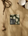 Geometry’s Paw Prints collection on a wooden floor: a brown leash, two white bowls on a stand, an ultra-absorbent Geometry Paw Prints towel with white spots, a black washcloth, and a chew bone.