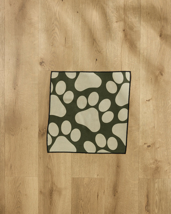 The Geometry Paw Prints mat, featuring large cream paw prints on green, sits on a light wood floor—perfect for catching messes after using an ultra-absorbent dog towel.