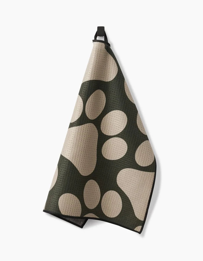 Geometrys Paw Prints kitchen towel features ultra-absorbent fabric in beige and dark green with an abstract oval pattern, shown hanging from a black loop on a plain white background.