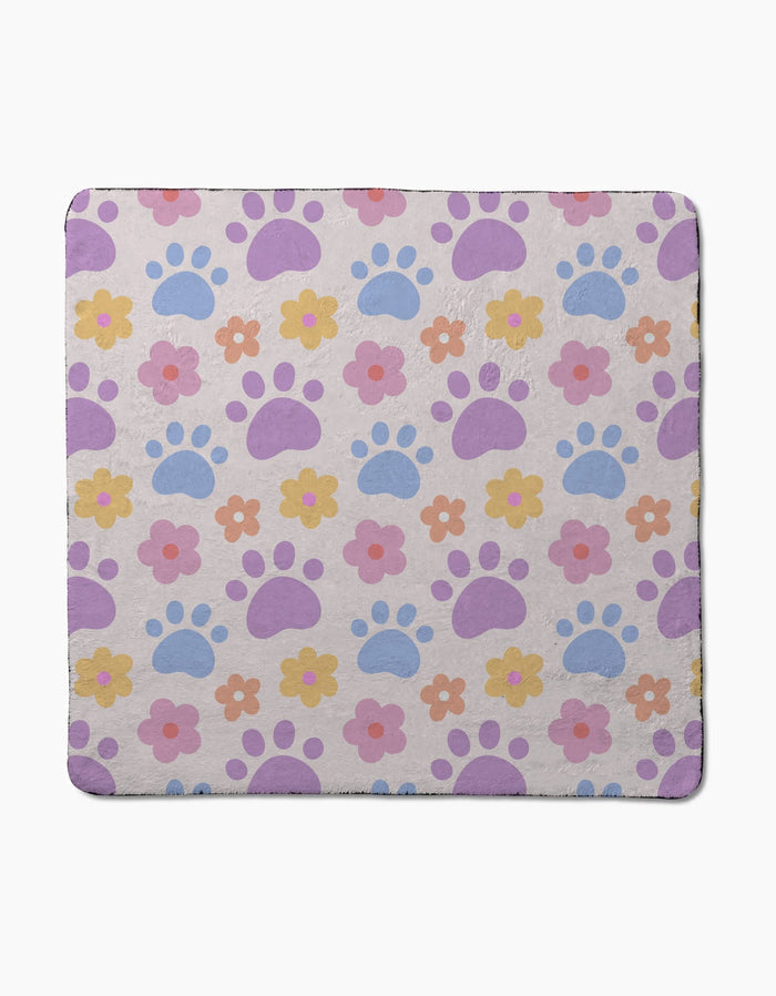 The Paws and Daisies dog towel by Letters By Shells showcases a pastel print of purple and blue paw prints with pink, yellow, and orange flowers on a light beige background.