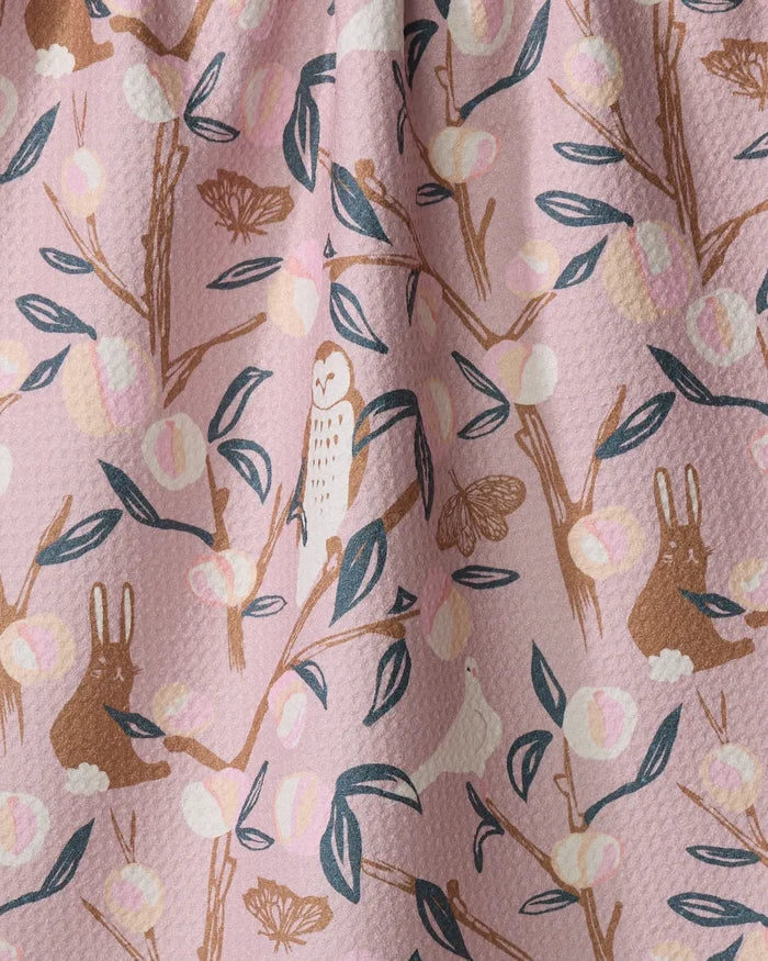 The "Peach Tree" kitchen tea towel by Elisabeth Wing features brown rabbits, owls, moths, leafy branches, and pink, cream, and gold fruits on a light pink background—an eco-friendly way to brighten any space.