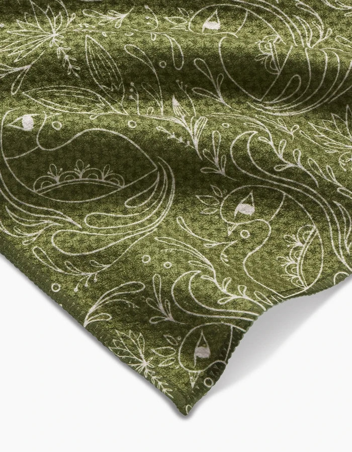 The Peacock Blooms Bar Towel by Amita Nair features green Geoweave fabric adorned with a white, whimsical bird and floral pattern. A folded corner showcases its soft texture and intricate design of stylized birds, leaves, and branches.