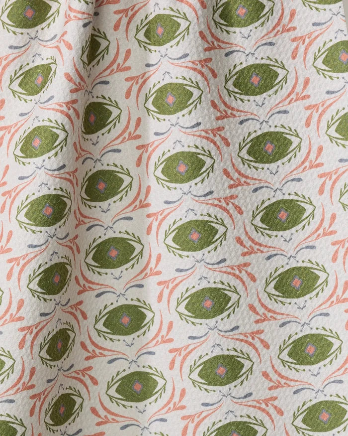 The "Peacock Eyes" tea towel by Amita Nair features green ovals with pink centers, bordered by pink and blue leafy accents on a cream background. Eco-friendly and slightly textured, it drapes softly—ideal for stylish kitchens.
