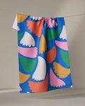 The Peacock Flowers tea towel by Jennifer Bouron features abstract wing-like shapes in pink, orange, yellow, green, and white on a blue background and hangs on a clothesline against a neutral wall.