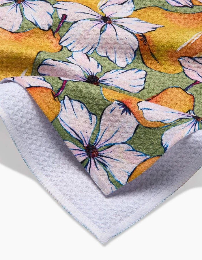 A close-up of the Pears Petals tea towel by Maayan Alper-Swan, featuring white flowers with purple accents on a green and yellow background, printed on one side and backed by white textured Geoweave fabric.