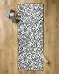 The Pebbles yoga mat by Marina Solodka, featuring a black and white pebble pattern, lies on a wooden floor with a non-slip towel. Two gray yoga blocks are at the top left, and a beige water bottle sits at the bottom right.
