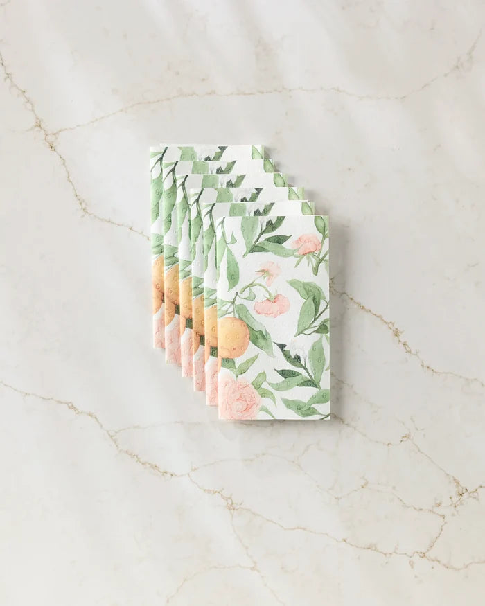 Six eco-friendly "Peony For Your Thoughts" kitchen towels by Aubrey Fairchild, featuring a floral and orange fruit pattern, are neatly stacked on a light marble surface.