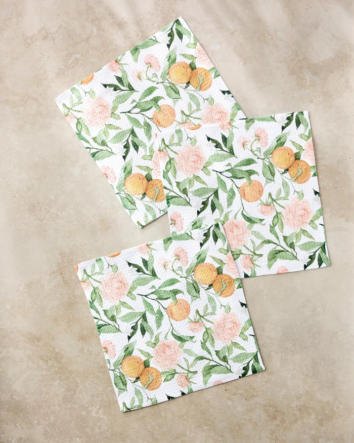 Three “Peony For Your Thoughts” square napkins by Aubrey Fairchild, crafted from recycled materials, display green leaves, peach flowers, and small orange fruits. They’re beautifully arranged on a light beige marble surface.
