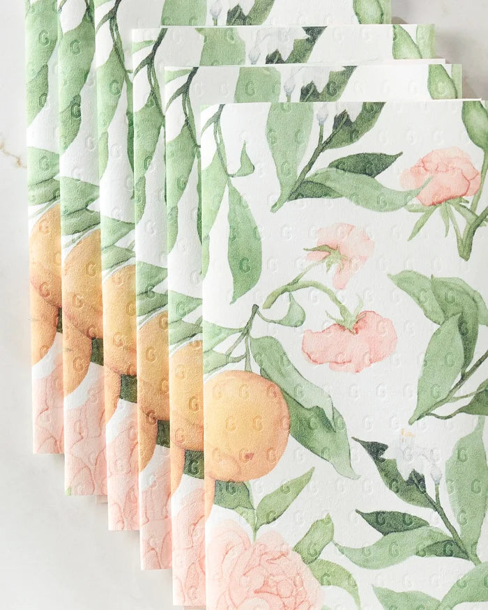 Several sheets of Aubrey Fairchild's "Peony For Your Thoughts" eco-friendly kitchen towels, showcasing green leaves, pink peonies, and orange fruits in a fanned-out display on a light surface.