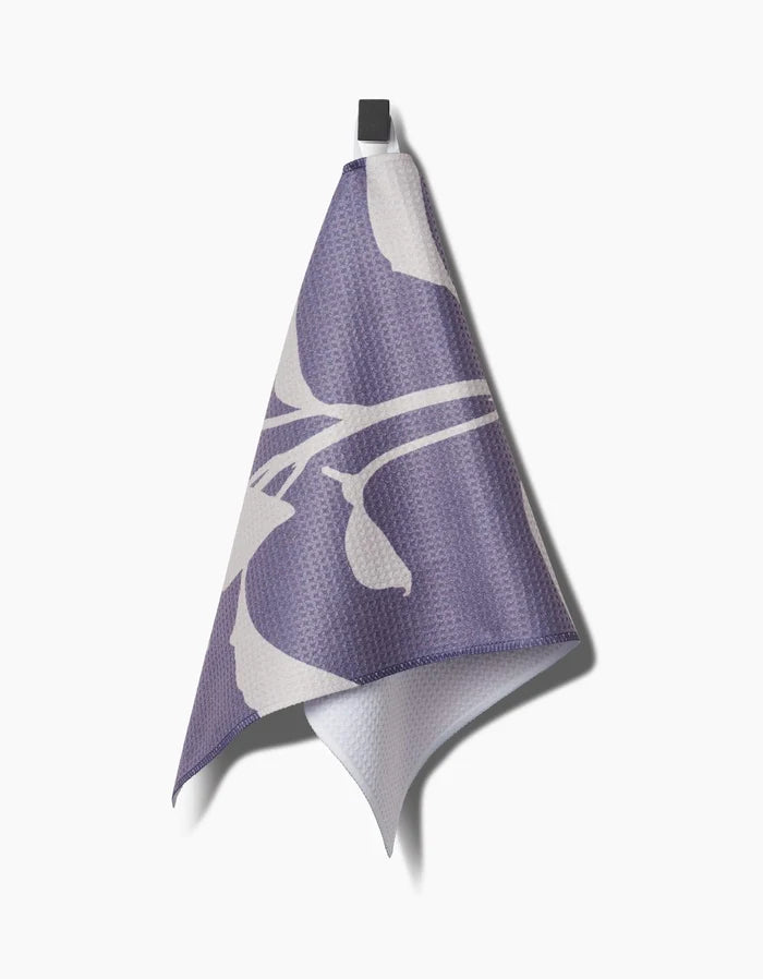A Pepper bar towel by PrintedPlant, featuring a bold purple and white abstract floral design made from recycled materials, hangs on a black hook against a plain white background.