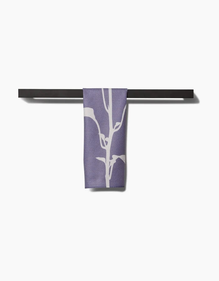 A PrintedPlant Pepper Bar Towel in purple with a white botanical design, made from Geoweave fabric, hangs over a black bar set against a plain white background.