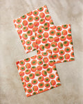 Three paper napkins by Katie O'Shea, featuring a repeating persimmons pattern, are arranged on a light beige marble surface, adding a vibrant touch reminiscent of an absorbent dishcloths set.