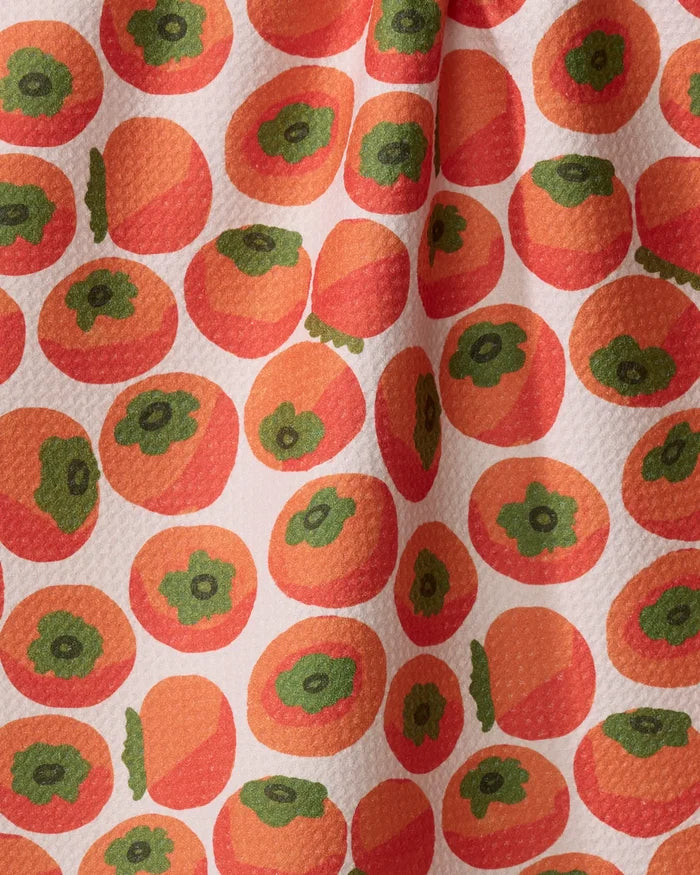 The "Persimmons" tea towel by Katie O'Shea features a repeating pattern of orange persimmons with green tops on a light, textured background. Some fruits overlap and soft fabric folds create gentle shadows.