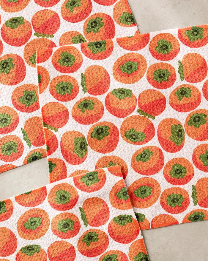 Several rectangular Persimmons dishcloths by Katie O'Shea, featuring a repeating orange and green persimmon pattern, are displayed overlapping on a light background.