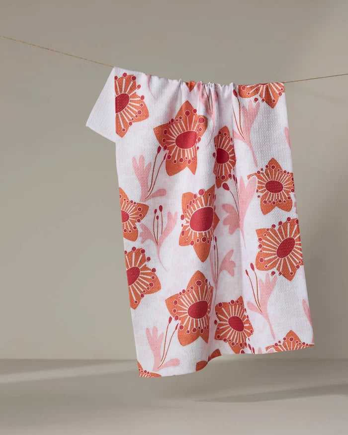 The Petal Paradise recycled kitchen towel by Whitney Urquhart, featuring a red and pink floral pattern, hangs on a clothesline against a neutral background.