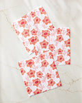 Three white Petal Paradise dishtowels by Whitney Urquhart, each featuring a red and orange floral pattern with stylized flowers and leaves, are displayed on a light marble-like surface.