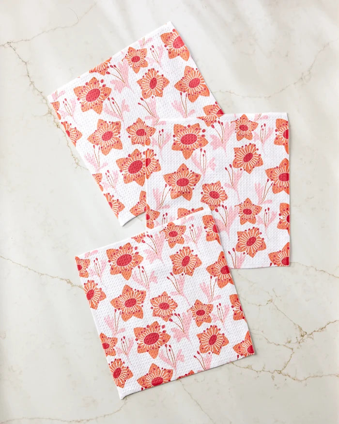 Three white Petal Paradise dishtowels by Whitney Urquhart, each featuring a red and orange floral pattern with stylized flowers and leaves, are displayed on a light marble-like surface.