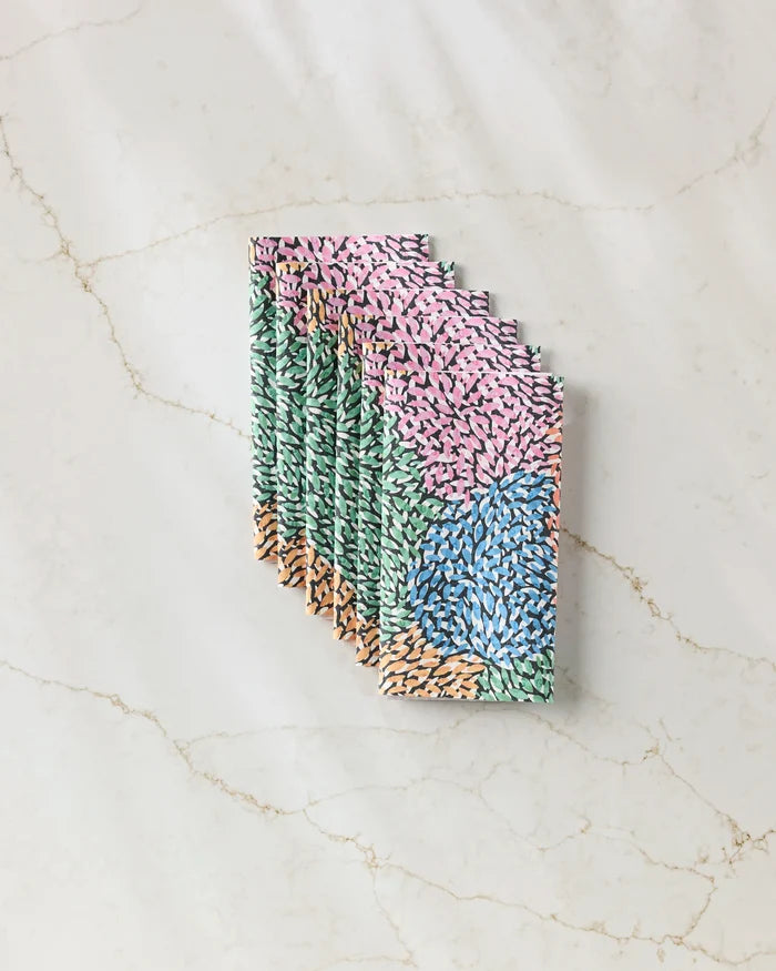 A stack of Petal Pen chocolate bars by Rachel Parker, wrapped in pink, green, blue, and orange abstract designs with small brushstroke shapes, rests on a faintly veined marble surface.