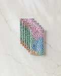 A stack of Petal Pen chocolate bars by Rachel Parker, wrapped in pink, green, blue, and orange abstract designs with small brushstroke shapes, rests on a faintly veined marble surface.