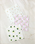 Three “Petals” paper napkins by Ania Zwara Art, made from recycled materials, are arranged on marble. The top has yellow and green dots, the middle shows pink flamingos, and the bottom is decorated with green avocados.