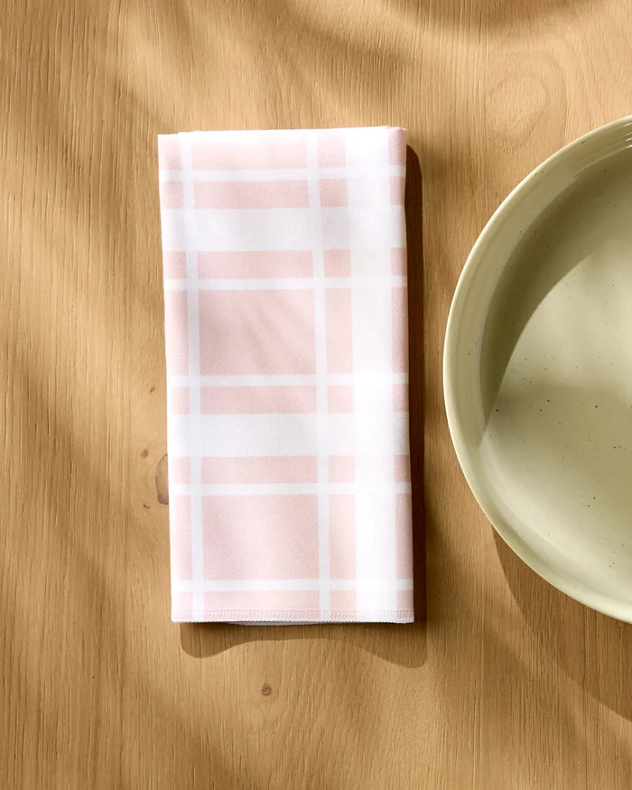 Aubrey Fairchild's Picnic Time Pink napkin, in pink and white plaid, rests folded on a light wooden table beside a cream ceramic plate, with sunlight casting gentle shadows across the scene.