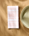 Aubrey Fairchild's Picnic Time Pink napkin, in pink and white plaid, rests folded on a light wooden table beside a cream ceramic plate, with sunlight casting gentle shadows across the scene.