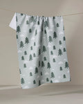 The Pine Forest kitchen tea towel by Coit Creative, featuring green pine trees and white clouds, is made from recycled materials and hangs on a clothesline against a neutral background.