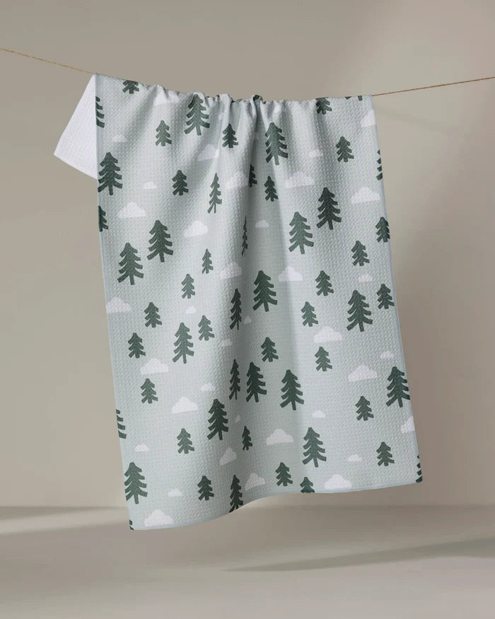 The Pine Forest kitchen tea towel by Coit Creative, featuring green pine trees and white clouds, is made from recycled materials and hangs on a clothesline against a neutral background.