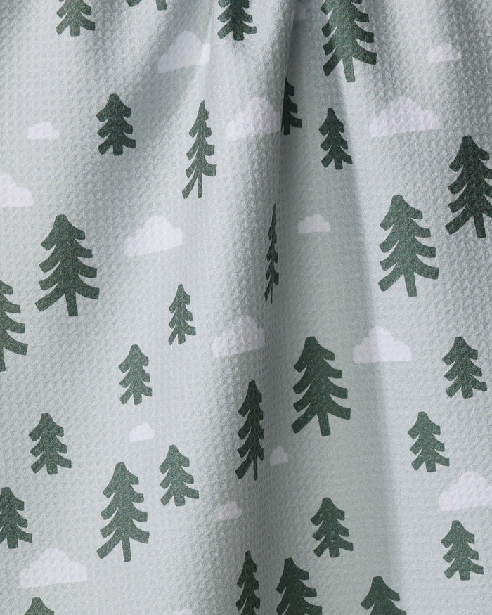 The Pine Forest tea towel by Coit Creative features a light gray textured fabric patterned with dark green pine trees and white clouds. Made from recycled materials, it's gently gathered at the top for soft folds.
