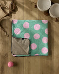 A plush dog towel with large pink polka dots is neatly folded on a wooden floor beside Geometry's Pink Ball, a brown leash, two white bowls, and several brown chew toys.