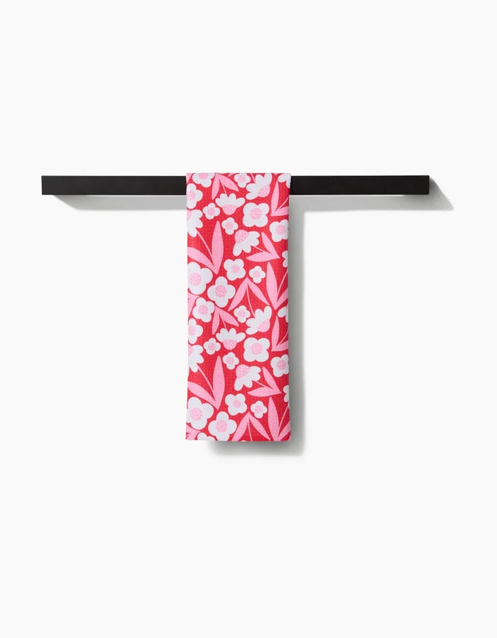 The Pink Blooms tea towel by Charlie Rowan Designs, featuring a white floral print on vibrant pink and made from quick-drying recycled materials, is displayed neatly draped over a sleek black rack against a plain white background.