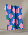 The Pink Lemons tea towel by Jennifer Bouron features large pink lemons with green leaves on a blue background and dries quickly.
